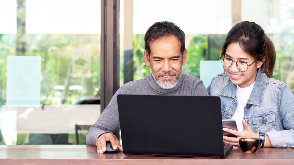 A smiling man and woman sit together at a table, sharing a laptop. The woman holds a phone. Bright, cheerful atmosphere with a window view.