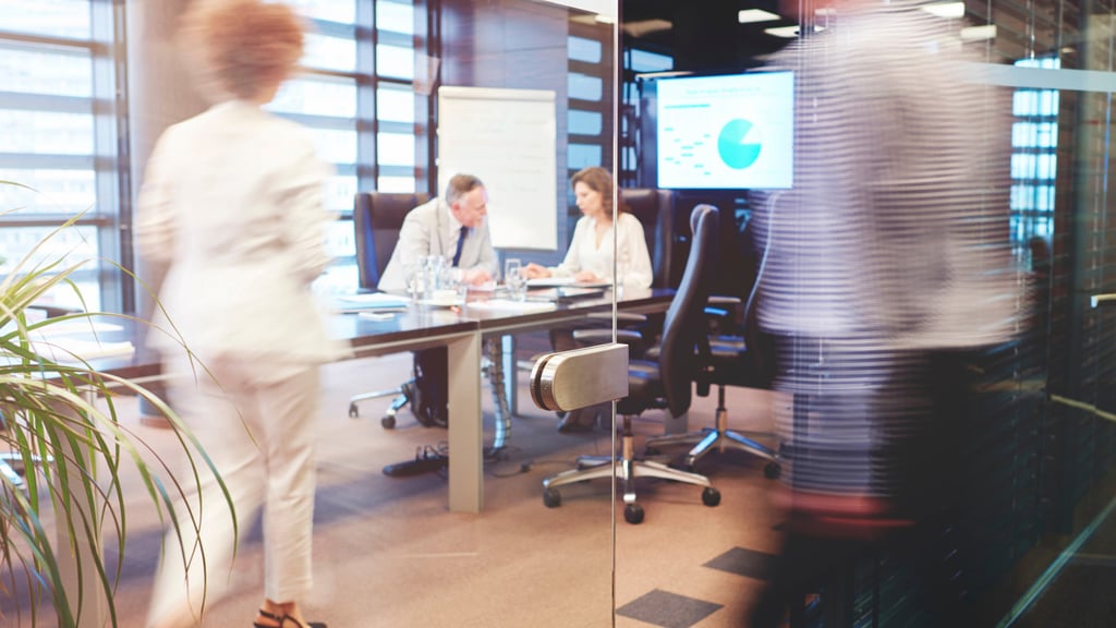 A modern office with two people in a meeting at a table, visible through glass walls. Motion-blurred figures walk by, conveying a dynamic atmosphere.