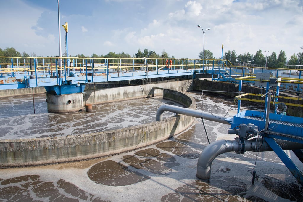 Wastewater treatment plant with large circular basins, aeration equipment, and blue railings. Cloudy sky and distant trees in the background.