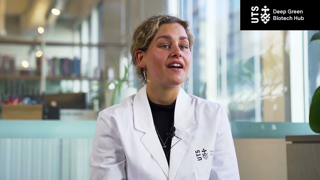 Person wearing a UTS lab coat seated indoors during an interview or presentation, with large windows and office shelving in the background. The UTS Deep Green Biotech Hub logo appears in the top-right corner.