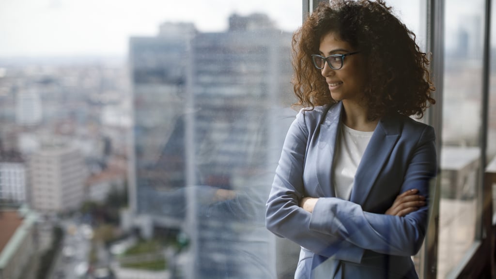 A professionally dressed women looks out a large glass window high up in an office building. She is wearing glasses and has her arms folded.