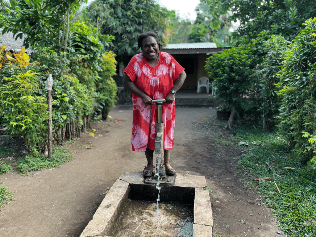 A person operates a hand water pump beside a rural pathway lined with dense greenery.