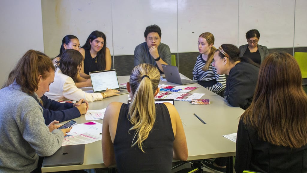 A group of students seated around a table in a classroom, collaborating using laptops and printed materials during a workshop discussion