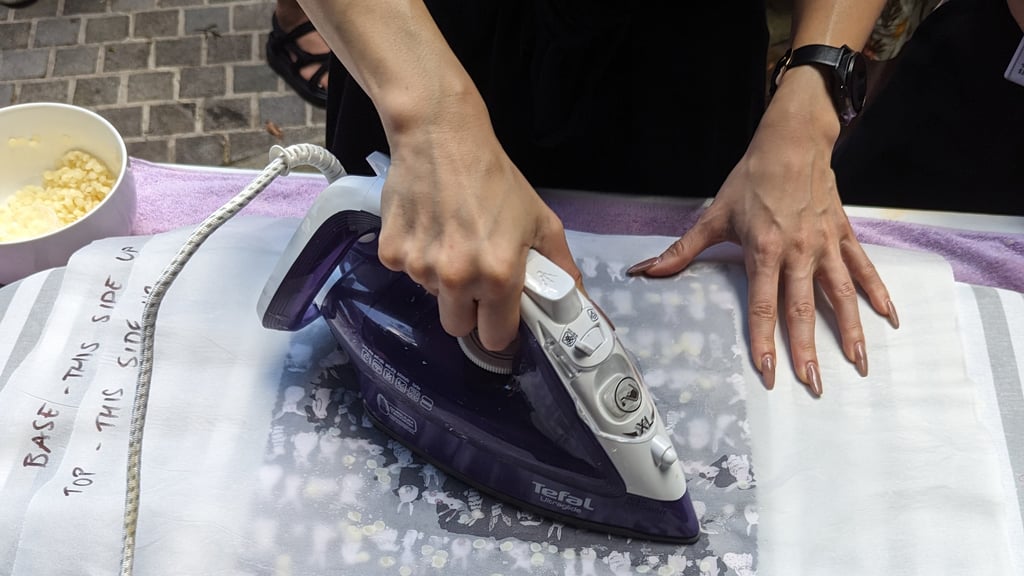 A person uses a purple iron on top of a sheet placed over a printed fabric. Their hands are focused, demonstrating a careful ironing process.