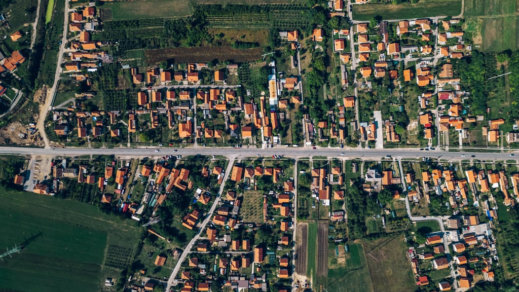 Aerial view of a suburban area with a straight road running horizontally. Red-roofed houses are neatly arranged amidst green fields and trees.