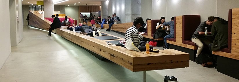 Students studying, collaborating and relaxing in a modern university corridor with long timber seating, booth-style study areas and open communal spaces.