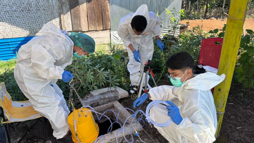 Three people wearing protective coveralls, gloves and face masks collect samples from an open well using scientific equipment in an outdoor rural setting.