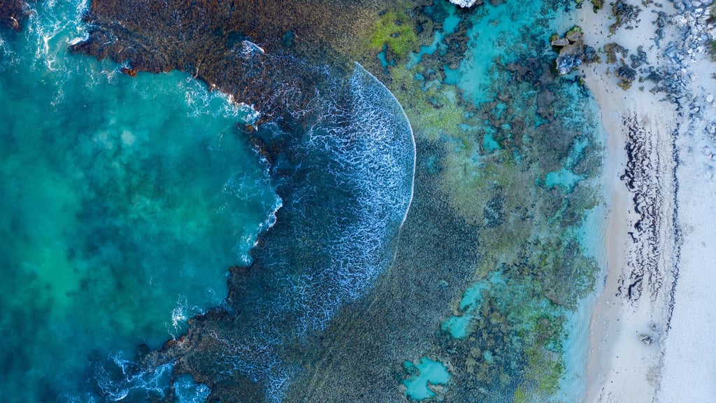 Aerial view of a vibrant shoreline with turquoise and deep blue waters meeting a sandy, rocky beach. Waves create a white arc on the sea, conveying tranquility.