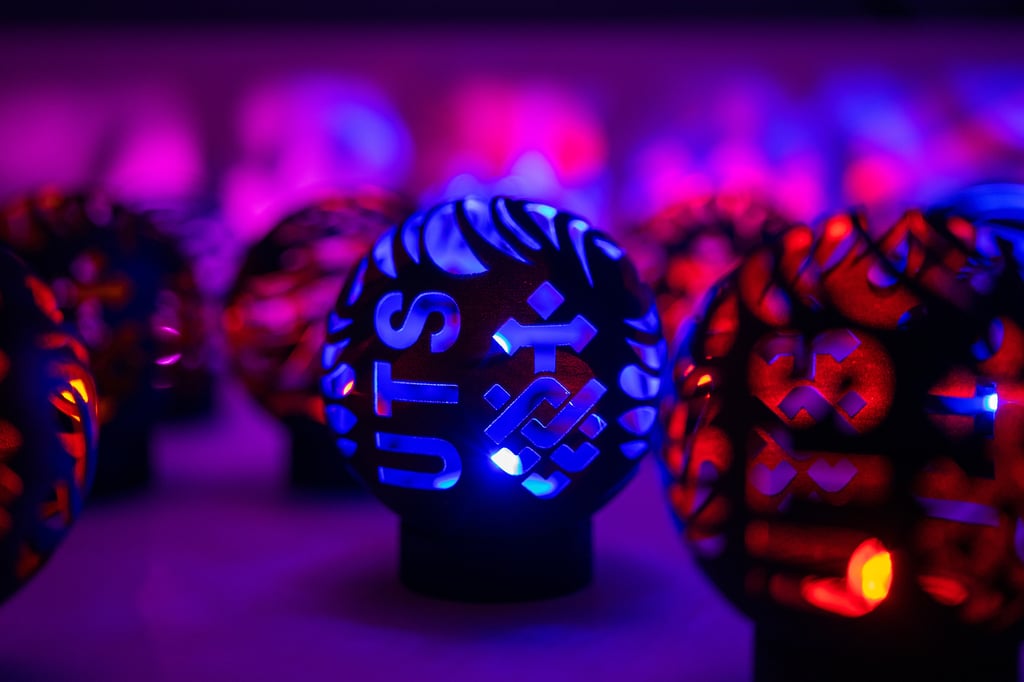 A spherical, black, hollow 3D printed part with blue LED lighting from within, featuring the UTS logo, among other unfocused parts in background.