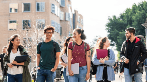 A diverse group of smiling students walks together on a sunny campus, carrying backpacks and notebooks. Modern buildings and trees are visible in the background.