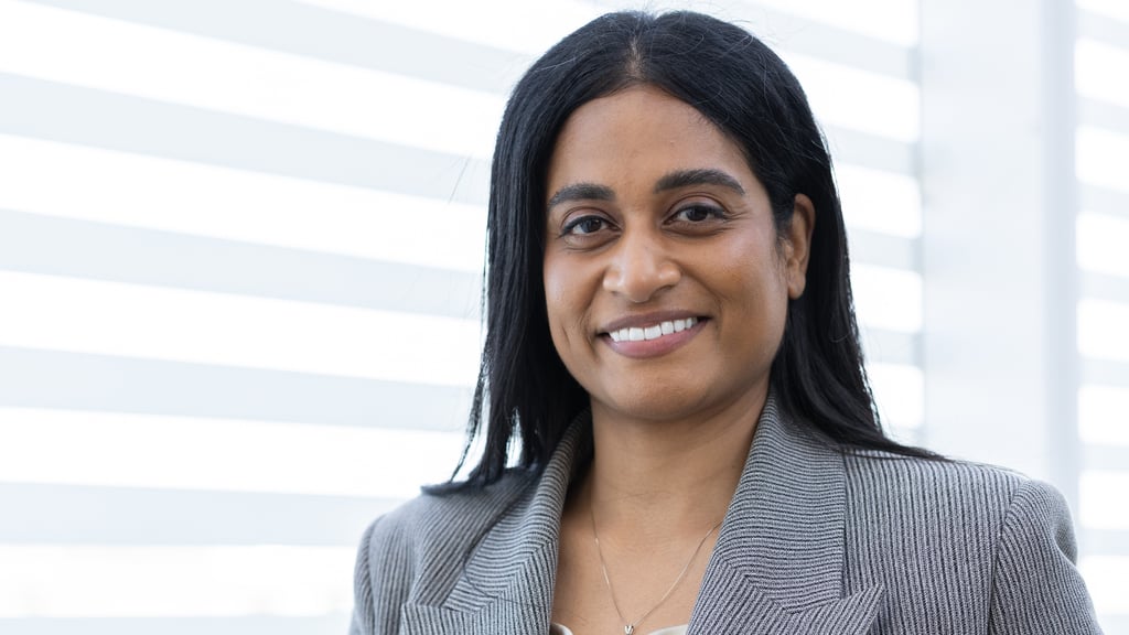 Ash wearing a grey blazer, smiling, standing in front of white blinds
