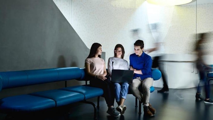 Three people sit closely on blue lounge chairs, focused on a laptop in a modern, dimly lit room with blurred figures walking by, conveying a sense of teamwork and activity.