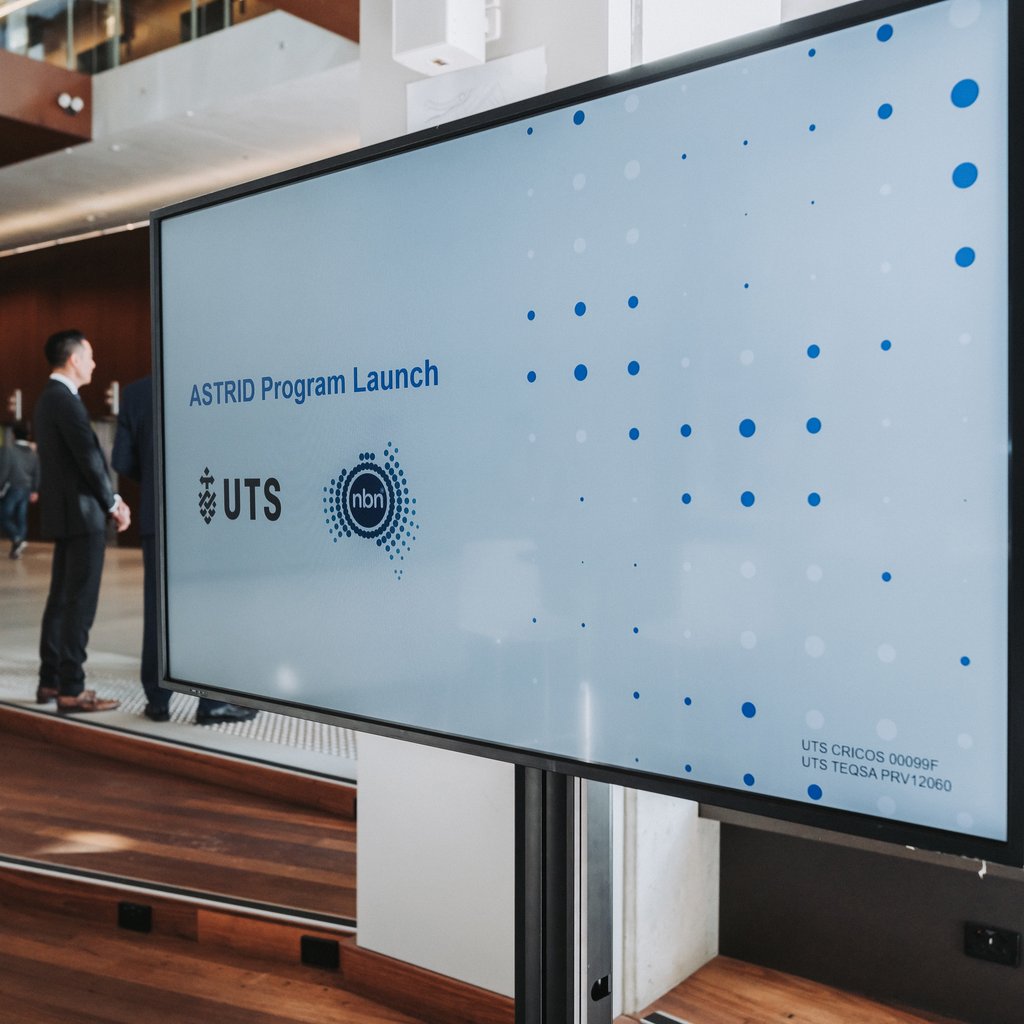 A large digital screen displays "ASTRID Program Launch" with UTS and NBN logos. A man in a suit stands nearby in a modern building setting.