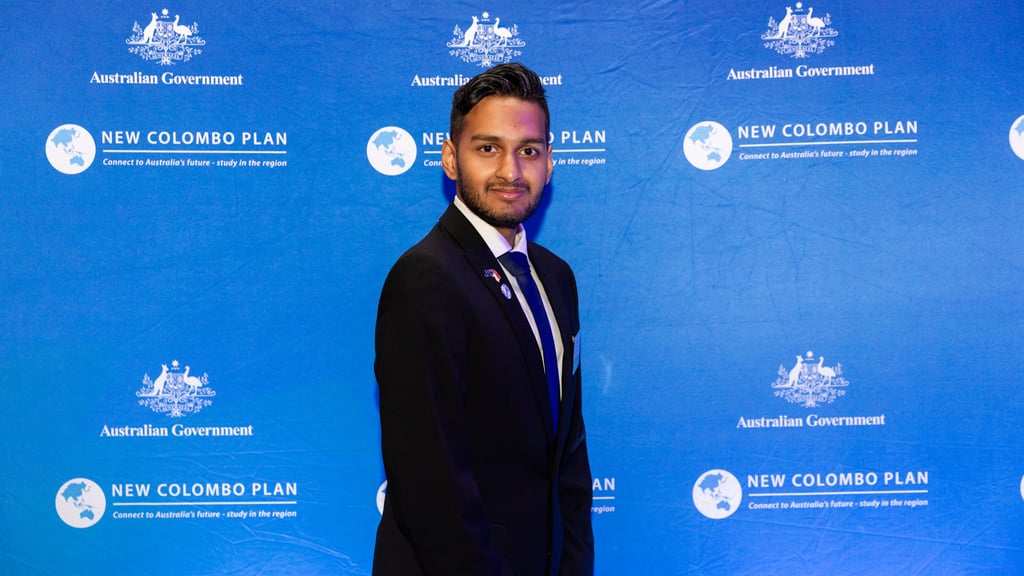 Sai received a 2024 scholarship from the New Colombo Plan Scholarships Program. Here he stands in front of a blue background to celebrate receiving the award