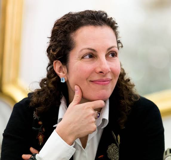Beth Simone Noveck is smiling at someone off-screen. She's dressed in black jacket with a white shirt and blue earrings.