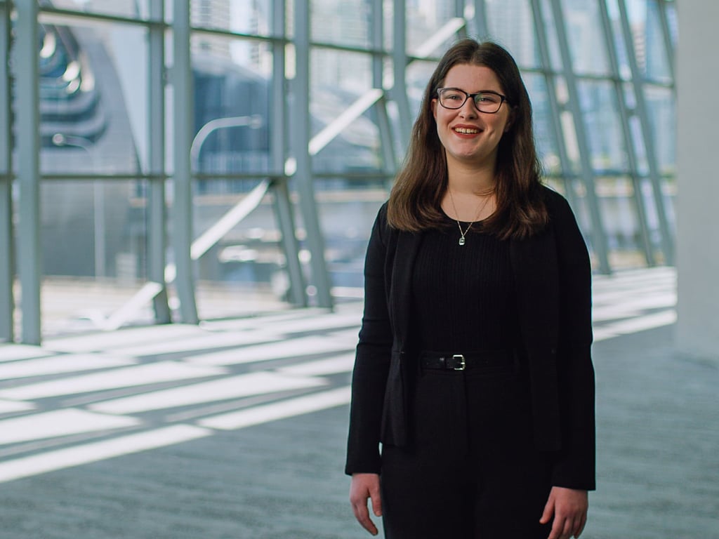 Emily Howe stands in the interior of the ICC Sydney's Convention Centre. She's wearing black pants and a blouse, smiling to the camera while in the background, tilted window panes show Sydney's Darling Harbour.