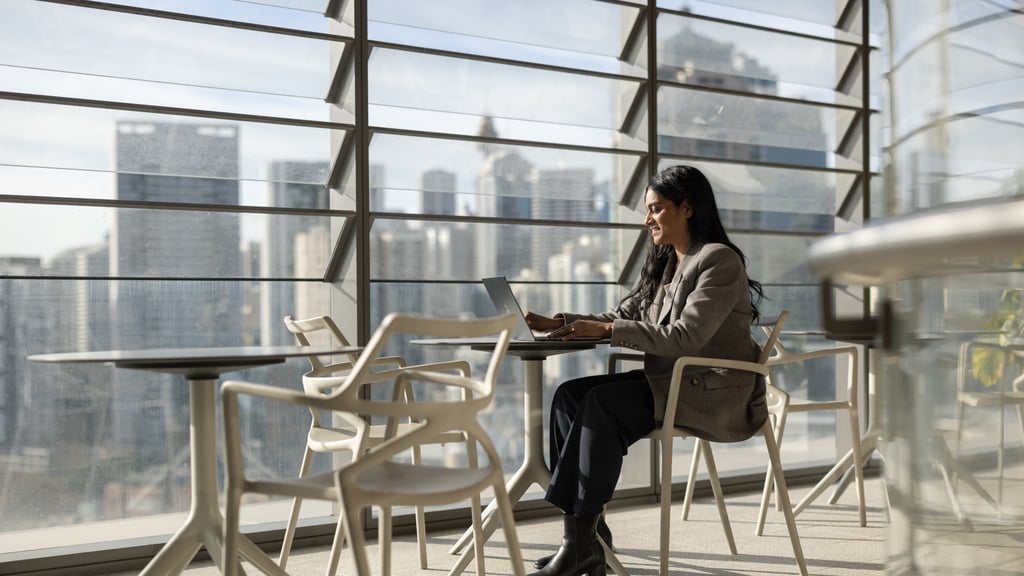 Ash sitting at a table working on her laptop with sun streaming in through the window, with the Sydney skyline visible