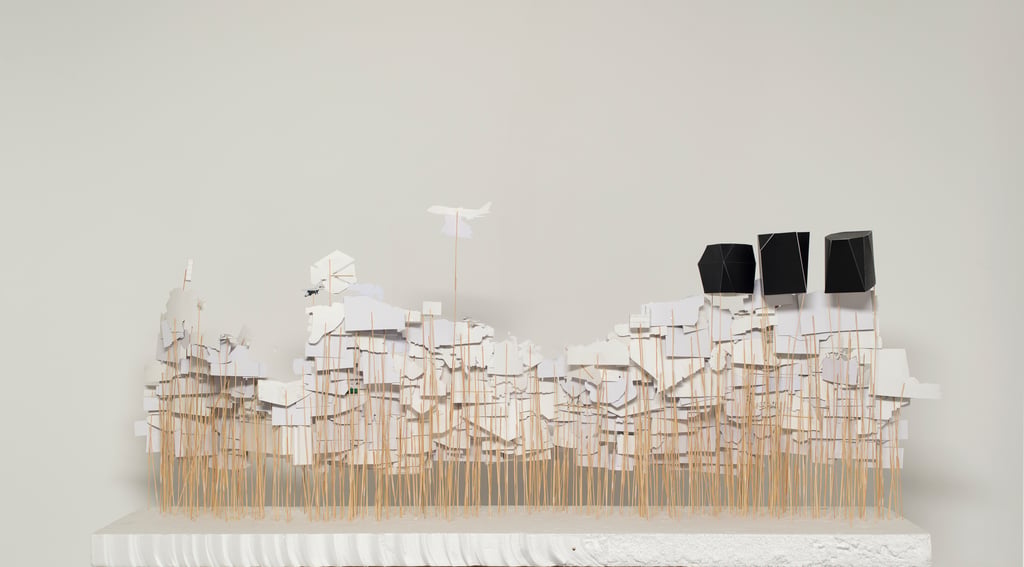 Art installation representing the city of Medellin consisting of pictures on paper and folded paper artefacts mounted on sticks of varying heights and embedded in a flat white base, seen from behind.