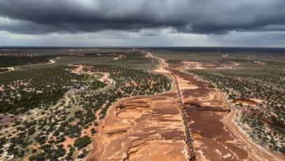 Aerial view of a wide, dry red desert landscape split by a dirt road. Sparse green vegetation surrounds. Dark storm clouds loom above, creating a dramatic tone.