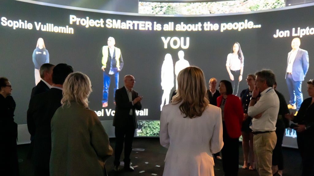 A group of people stand in a circular room with a large, curved screen displaying the text "Project SMaRTer is all about the people" and individual names. The atmosphere is engaged and collaborative.