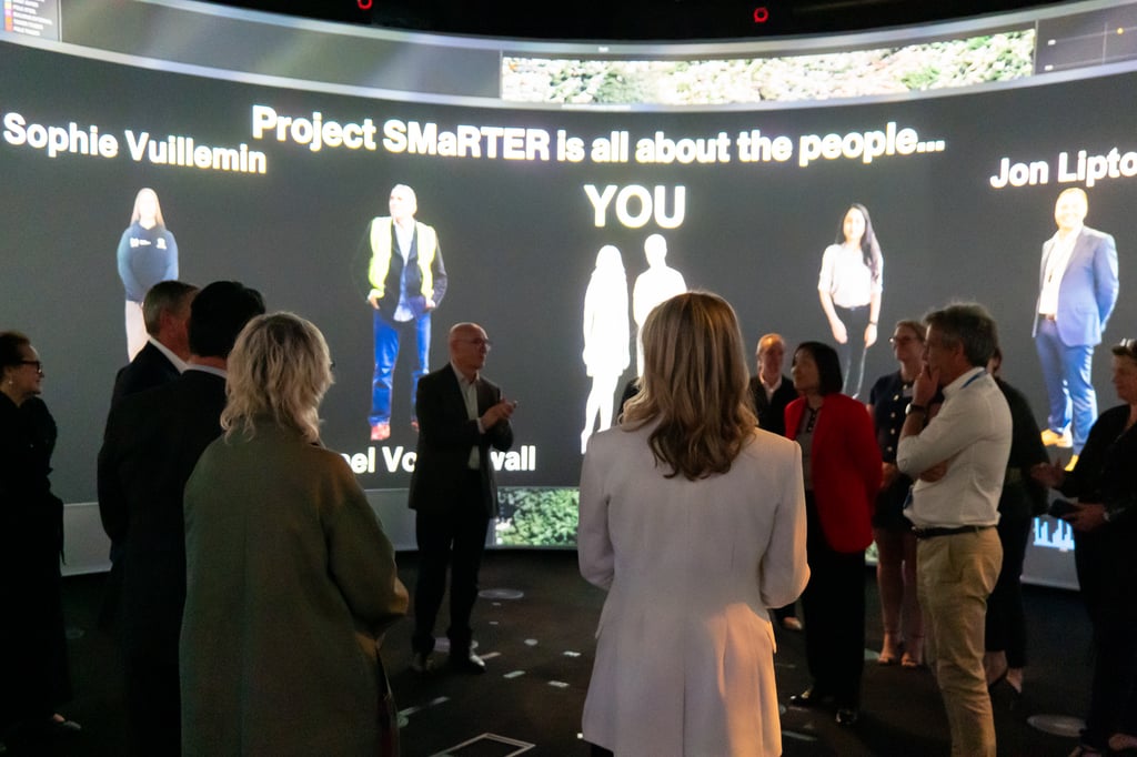A group of people stand in a circular room with a large, curved screen displaying the text "Project SMaRTer is all about the people" and individual names. The atmosphere is engaged and collaborative.