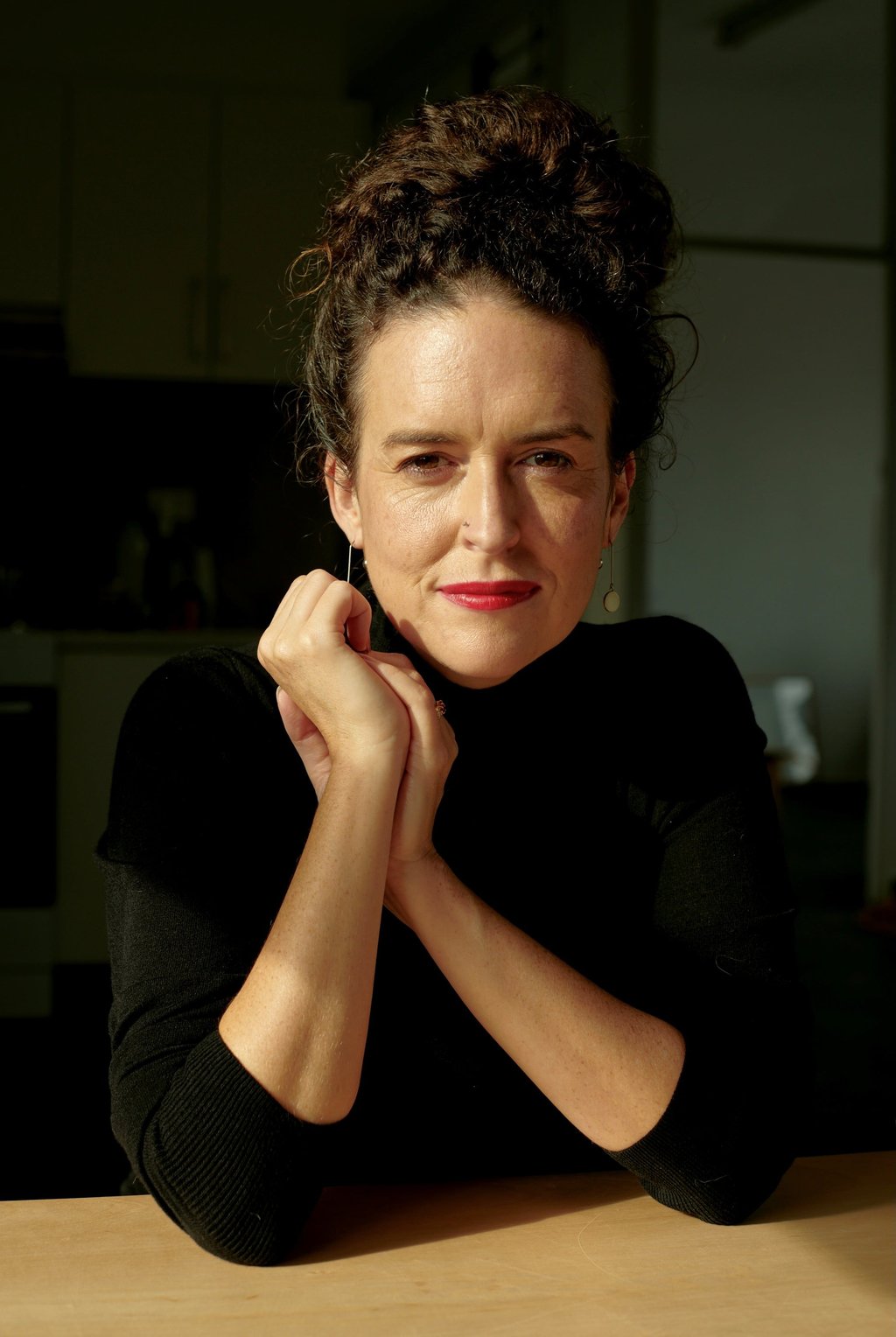 Jess Hill is seated indoors with her hands gently clasped under her chin, wearing a black long‑sleeve top. Soft, warm light highlights her arms and hair, with a dimly lit kitchen in the background.