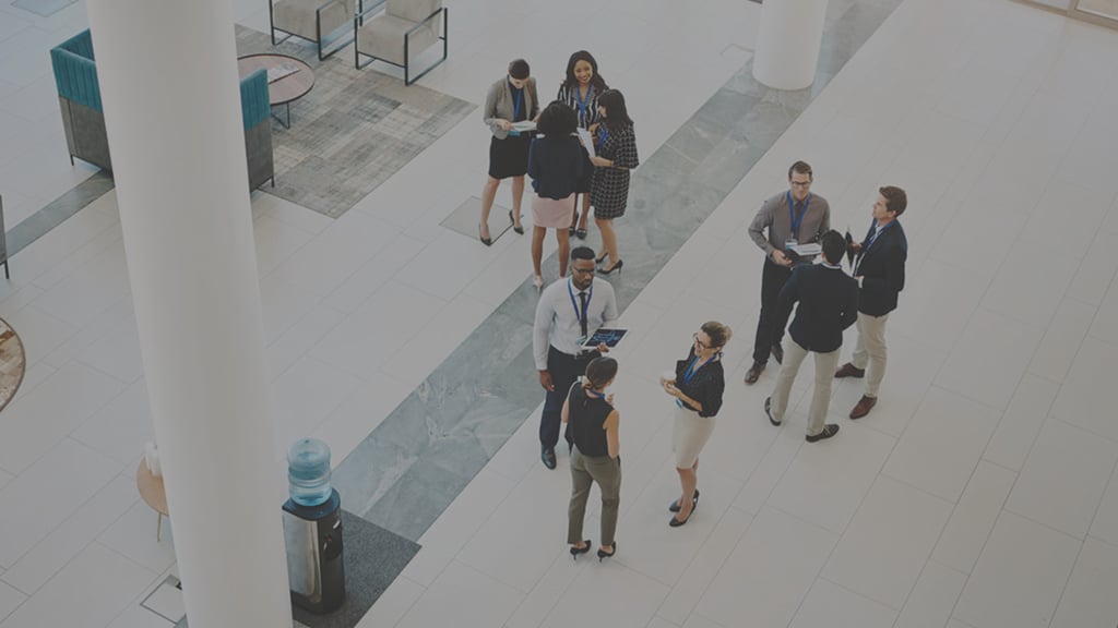 People gathered around and speaking together in a building foyer as seen from an atrium above.