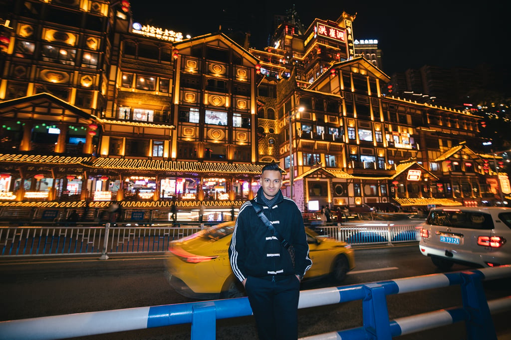 Sai stands in front of an entertainment precinct in Asia. It is night and the buildings and restaurants are lit up.