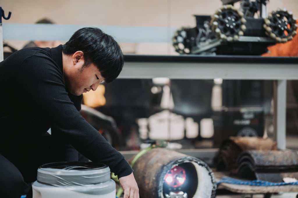 A person in a black shirt is focused on adjusting a cylindrical device with glowing lights in a workshop. A robotic vehicle is visible on a table behind them.