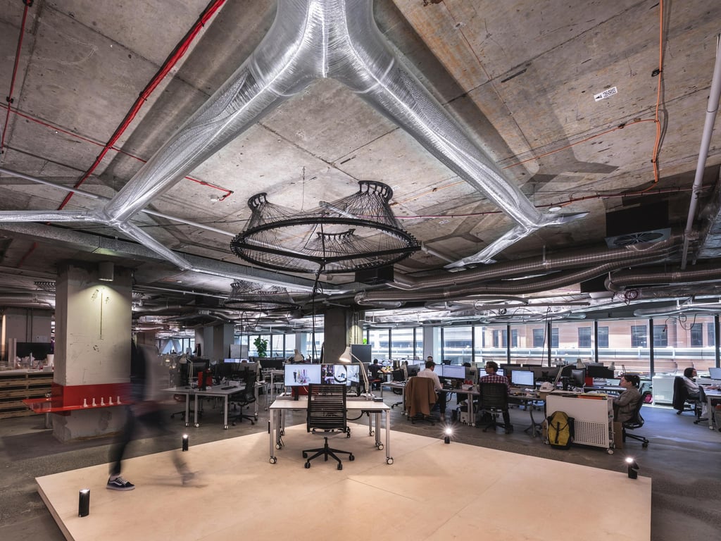 Open office with industrial ceiling, exposed ducts, and modern desks. People work at computers under bright lighting, creating a collaborative atmosphere.
