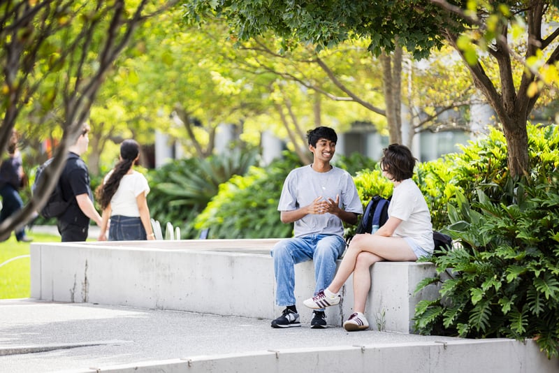 Two people sit and talk on a concrete bench in a leafy outdoor area at UTS, with other people walking along a path in the background.