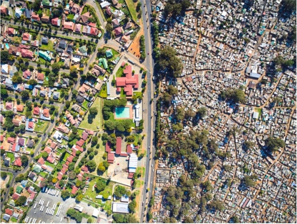 Aerial view showing a stark divide between a well-planned suburban neighborhood with large houses on the left and a densely packed settlement on the right, separated by a road.