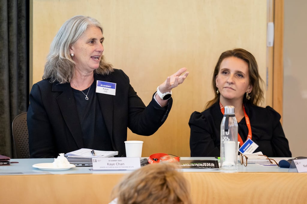 Dr. Kaye Chan sits at a table during a discussion, with one gesturing while speaking addresses summit attendees who cannot be seen within the image, notebooks, name cards and water bottles are arranged in front of her.