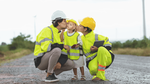 A child in a yellow safety vest and hard hat stands between two adults in reflective gear, sharing a loving moment on a rural road under an overcast sky.