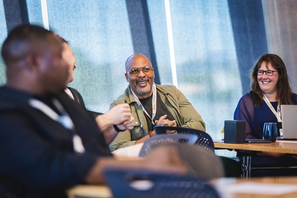 Participants seated around a meeting table during a collaborative workshop, with discussion taking place among attendees wearing conference lanyards.