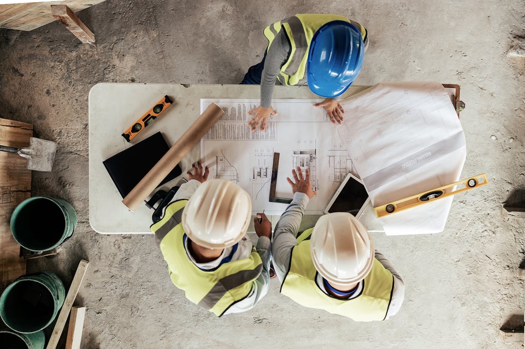 Three construction workers in safety vests and helmets reviewing building plans on a table. The scene conveys teamwork and planning at a construction site.