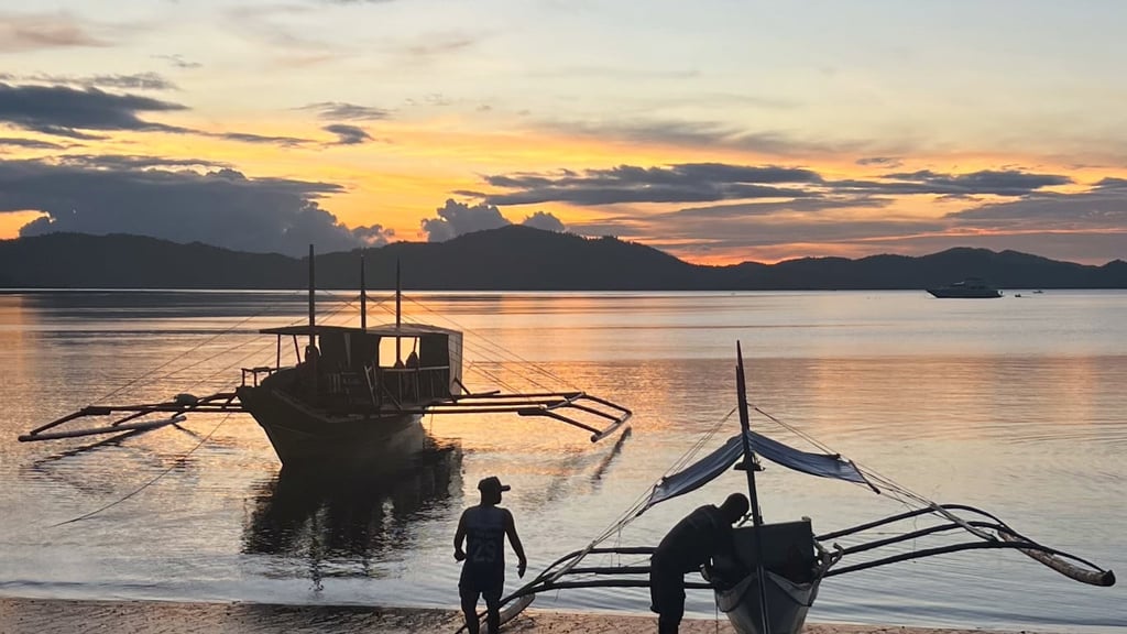 Sunset over calm waters with two silhouetted boats in the foreground and distant mountains. A tranquil scene with soft orange and blue hues.