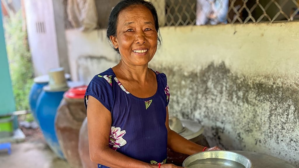 A woman in a blue floral dress smiles warmly while holding a metal bowl. She stands in front of a rustic wall with household items in the background.