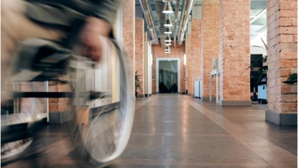 A person in a wheelchair moves through a modern hallway with exposed brick pillars and hanging lights, conveying motion and accessibility.