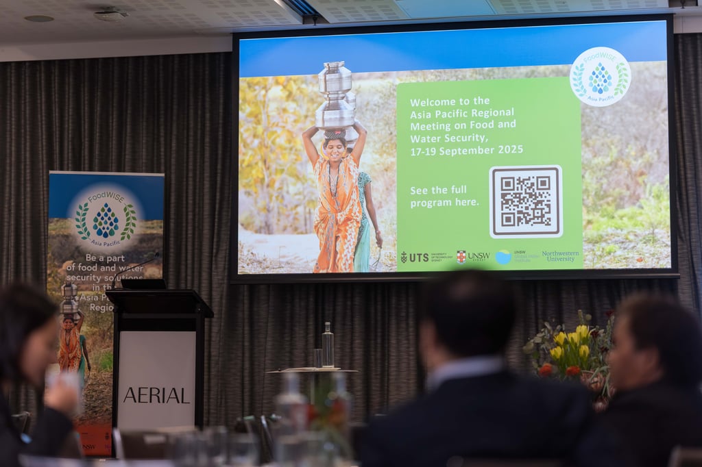 A conference room displays a large screen showing an announcement for the Asia Pacific Regional Meeting on Food and Water Security, dated September 17-19, 2025. The screen features an image of a person in traditional attire carrying water pots on their head. Attendees are seated at tables with floral decorations, creating a formal atmosphere.
