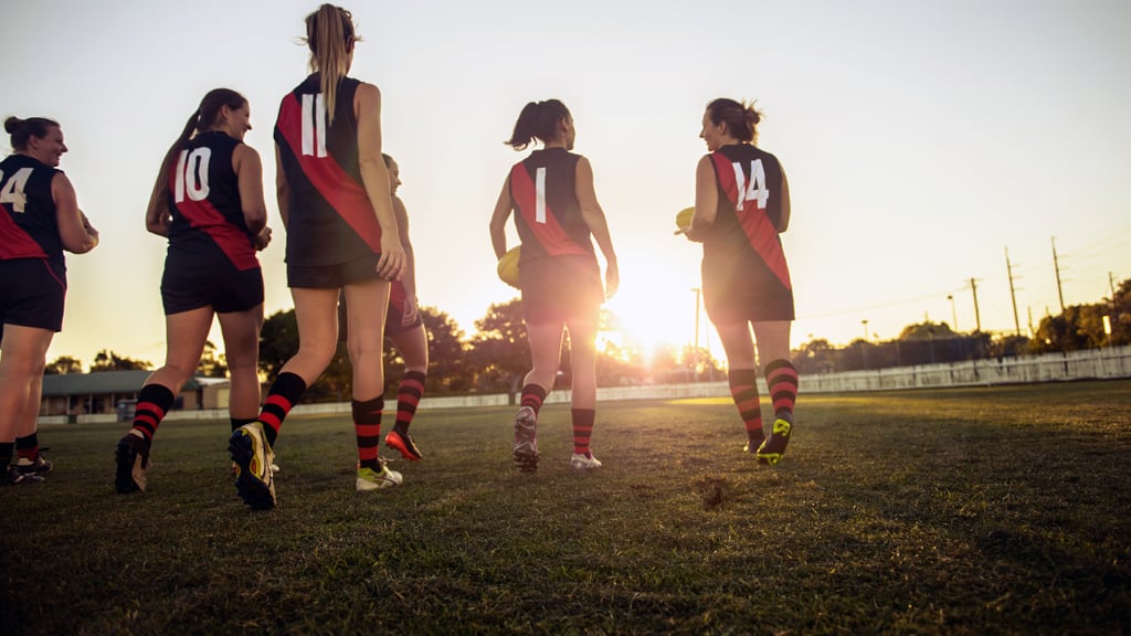 Six female Australian Rules football players run on to the training paddock as the sun sets behind them.
