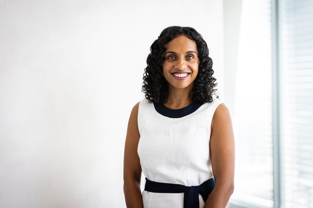 Ramona Vijeyarasa smiles. wearing a white dress with black trim. She stands in front of a white wall and a window with blinds