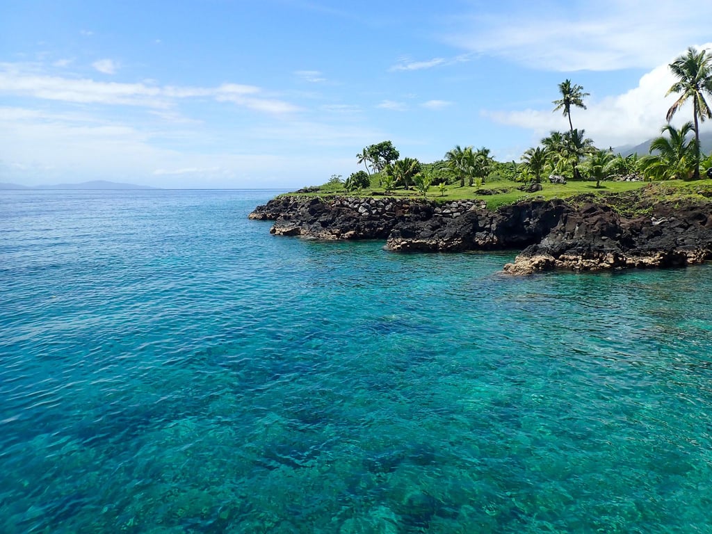 Clear turquoise ocean water meets rocky cliffs with lush greenery and palm trees against a bright blue sky, evoking a serene tropical paradise.