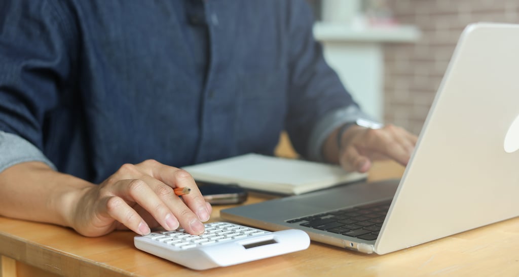 A person in a blue shirt works at a wooden desk using a laptop and a calculator, with a pen and notebook nearby, conveying focus and productivity.