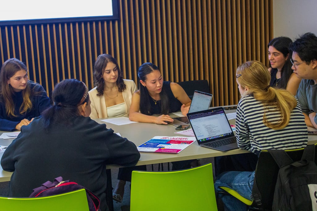 Students gathered around a table reviewing a laptop screen and printed handouts, with one participant gesturing while leading the discussion