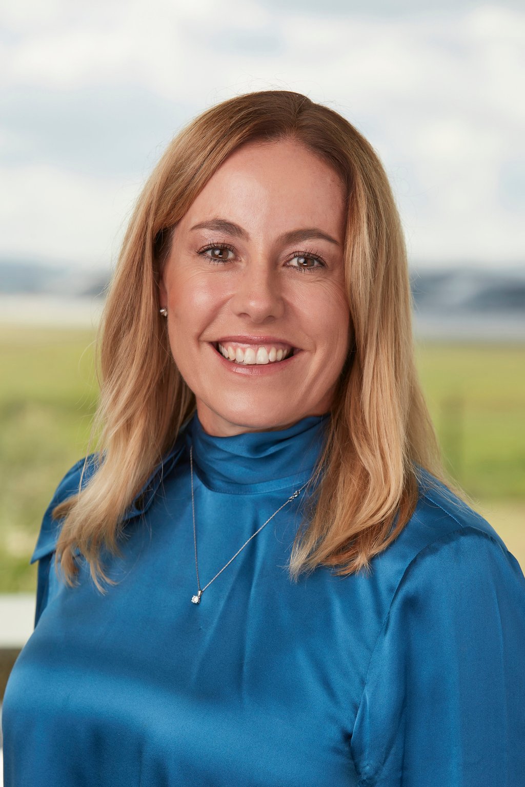 Portrait of Shelley Cole, she stands in the foreground smiling to camera. She is wearing a blue dress and has blonde hair. In the background is the Western Sydney International Airport.