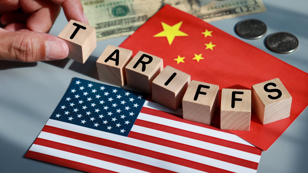 Wooden blocks spelling "TARIFFS" placed between the flags of the United States and China, with U.S. currency (dollar bills and coins) in the background