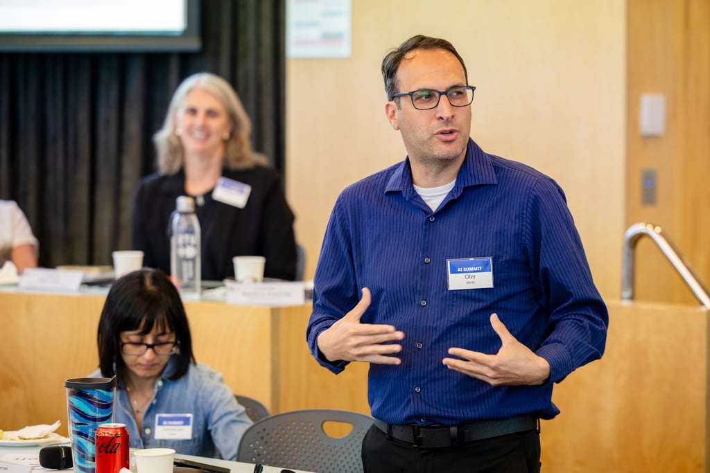 Associate Professor Ofer Mintz stands in the centre of the UTS Business School Oval Classroom and gestures while speaking during a workshop discussion, with seated attendees and refreshments visible around the table.