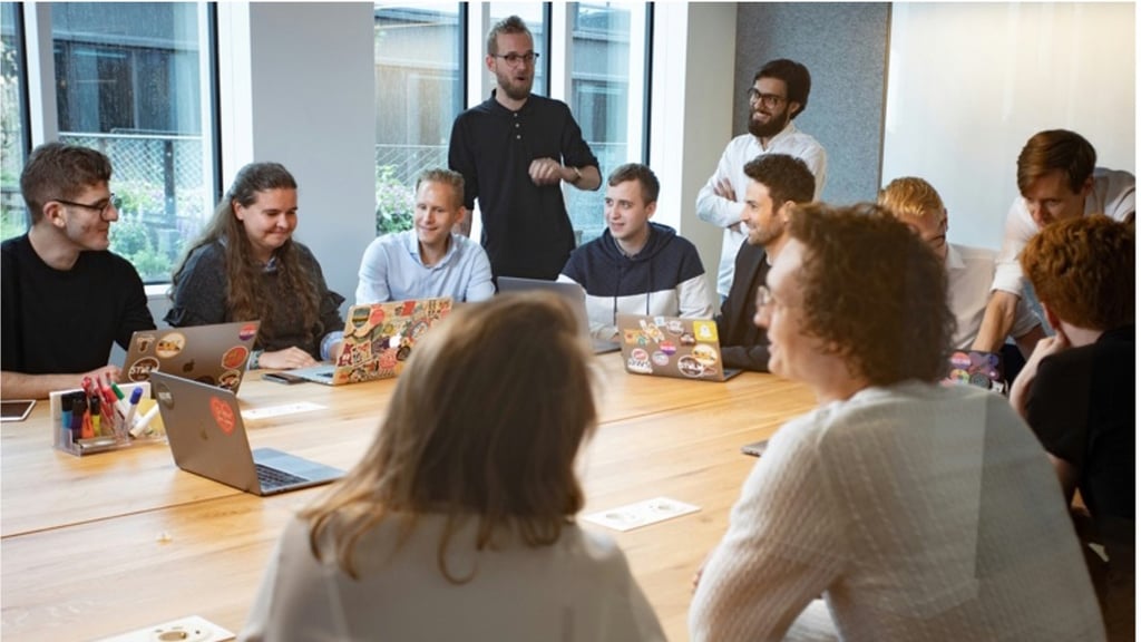 Group of young professionals around a conference table with laptops decorated with stickers, engaged in a lively discussion, smiling and focused.