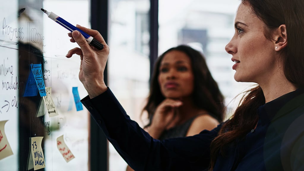 Women pointing to a whiteboard with a marker pen while another woman watches closely.
